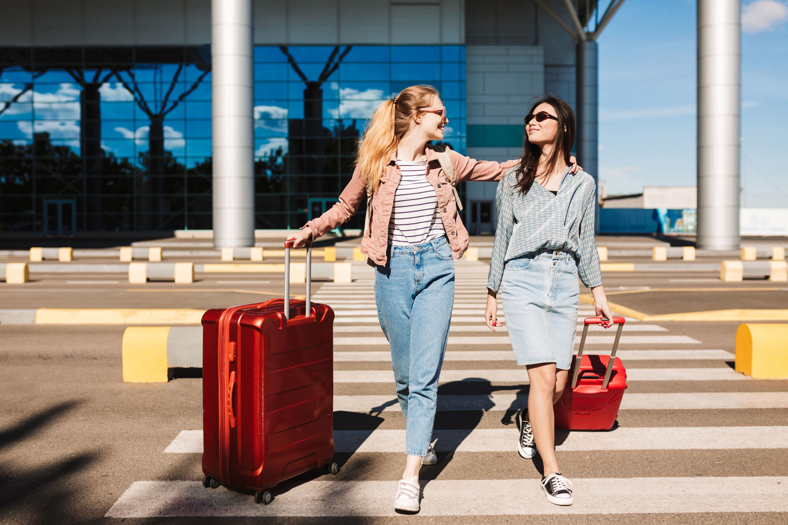 beautiful-stylish-girls-sunglasses-happily-walking-along-pedestrian-strip-with-red-suitcases-airport-background-scaled